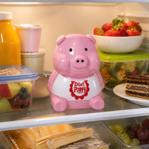 Diet Piggy koelkastalarm op een koelkastplank naast aardbeien, yoghurt, fruit en een stukje taart.