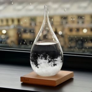 Een druppelvormig stormglas op een houten voet, geplaatst op een vensterbank voor een raam met regendruppels en een wazig stadsgezicht op de achtergrond.