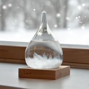 Een klein, druppelvormig stormglas op een houten voet, geplaatst op een vensterbank met een wazig, besneeuwd winterlandschap op de achtergrond.